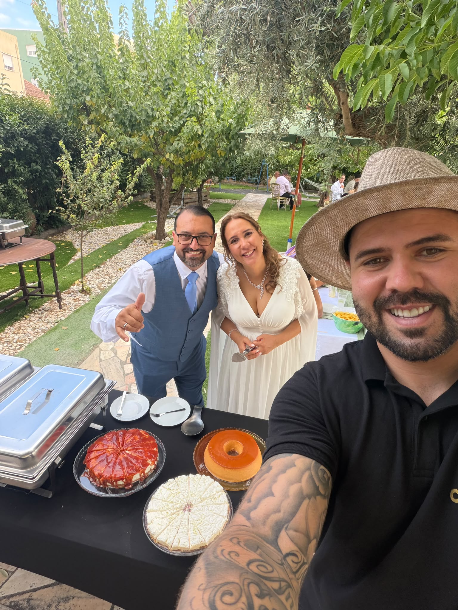 Caterer com chapéu a tirar selfie com os noivos junto à mesa de sobremesas decorada num jardim