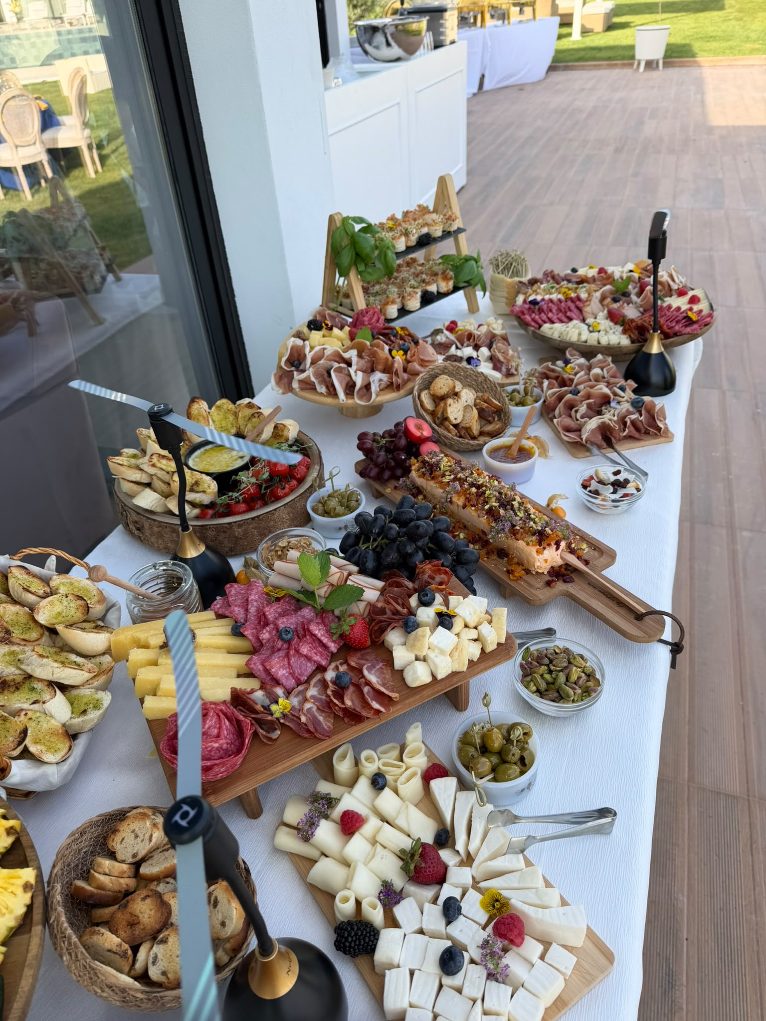 Mesa de charcutaria gourmet com queijos, enchidos, frutos, tostas e molhos num pátio ao ar livre