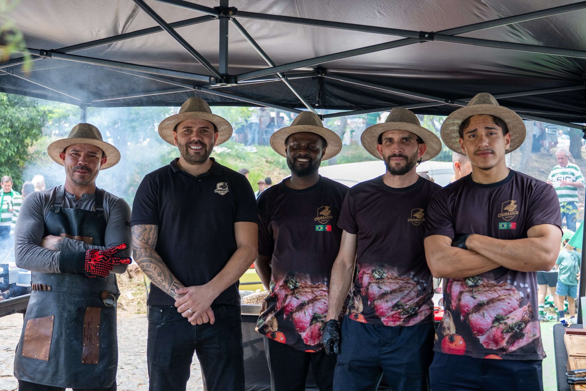 Equipa de cinco churrasqueiros do Churrasco da Elite com chapéus de palha e t-shirts da marca, juntos sob tenda de evento