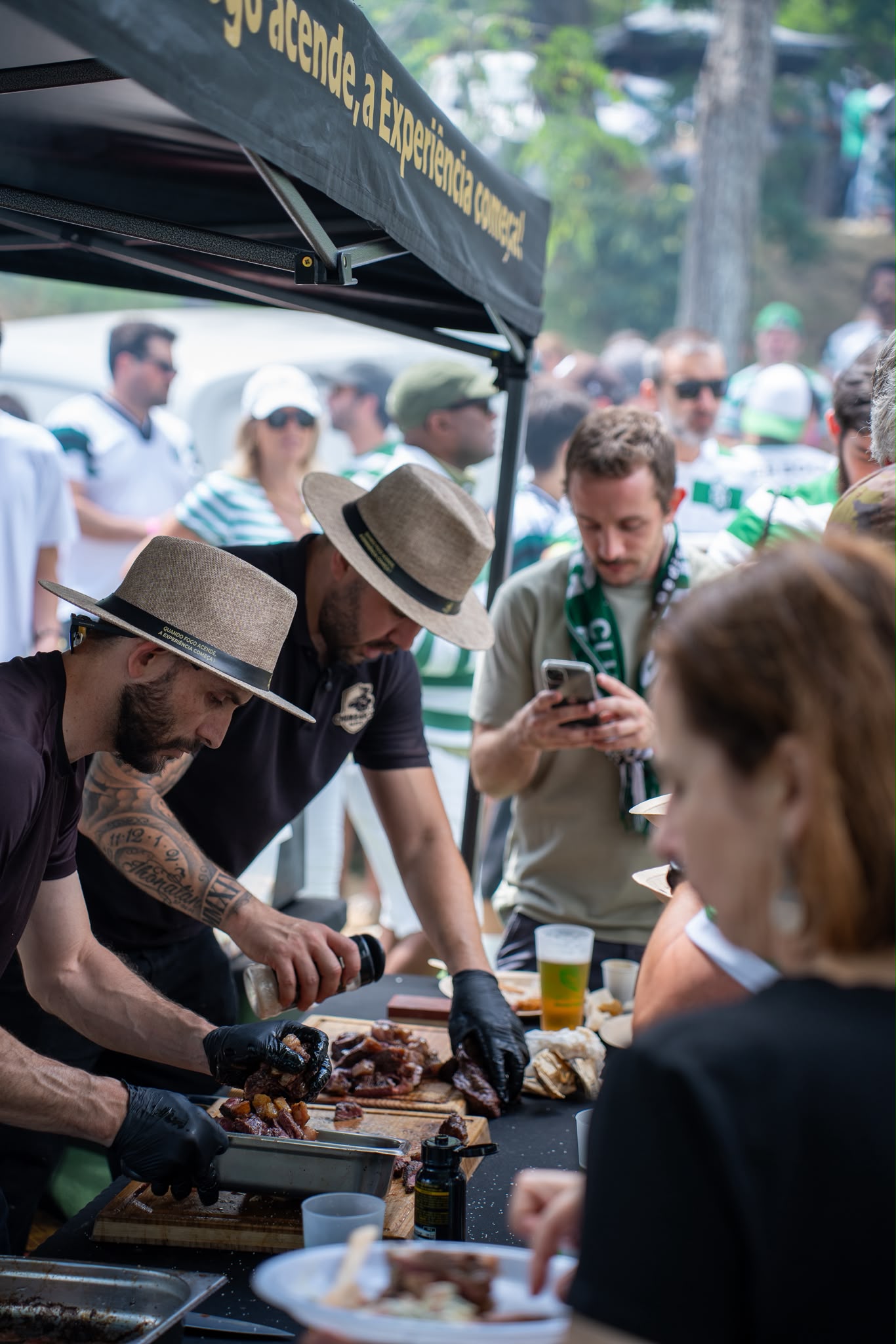 Dois chefs do Churrasco da Elite com chapéus de palha a preparar carnes grelhadas num evento ao ar livre