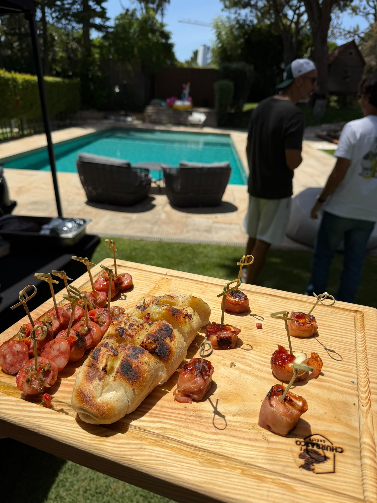 Tábua de aperitivos com salsichas grelhadas, wraps e pão de alho junto à piscina num evento ao ar livre